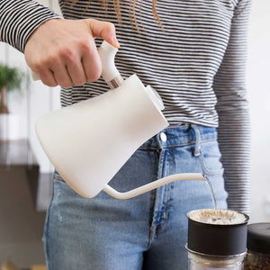 Ruka nalévá vodu z bílé rychlovarné konvice s úzkým hrdlem do kávového filtru, část EspressoGear, vše o kávě.